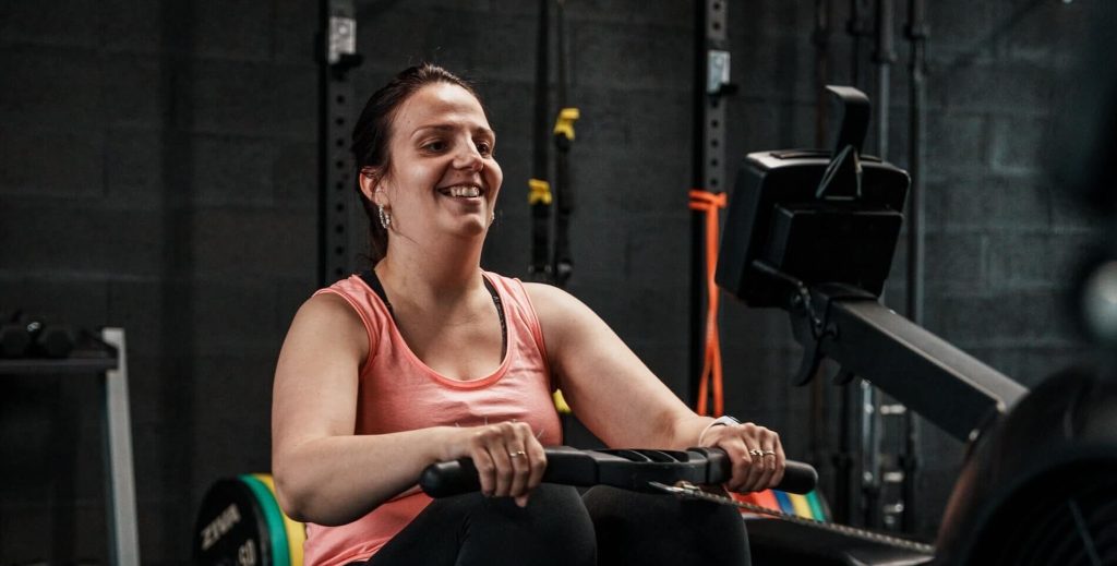 Femme souriante pendant une séance de rameur en fin de journée, illustrant le sport le soir à intensité modérée chez Ultimate Physical