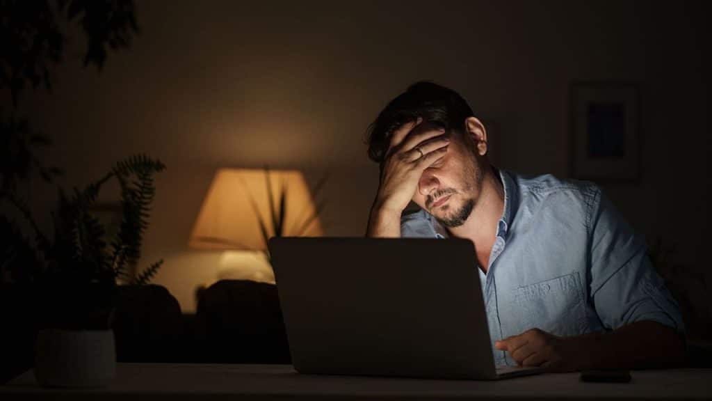 Douleur cervicale télétravail : homme travaillant tard le soir sur un ordinateur portable, la tête penchée en avant