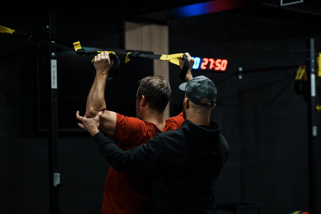 Douleur épaule musculation : coach sportif sécurisant le bras d'un client pendant un exercice au TRX bras au-dessus de la tête
