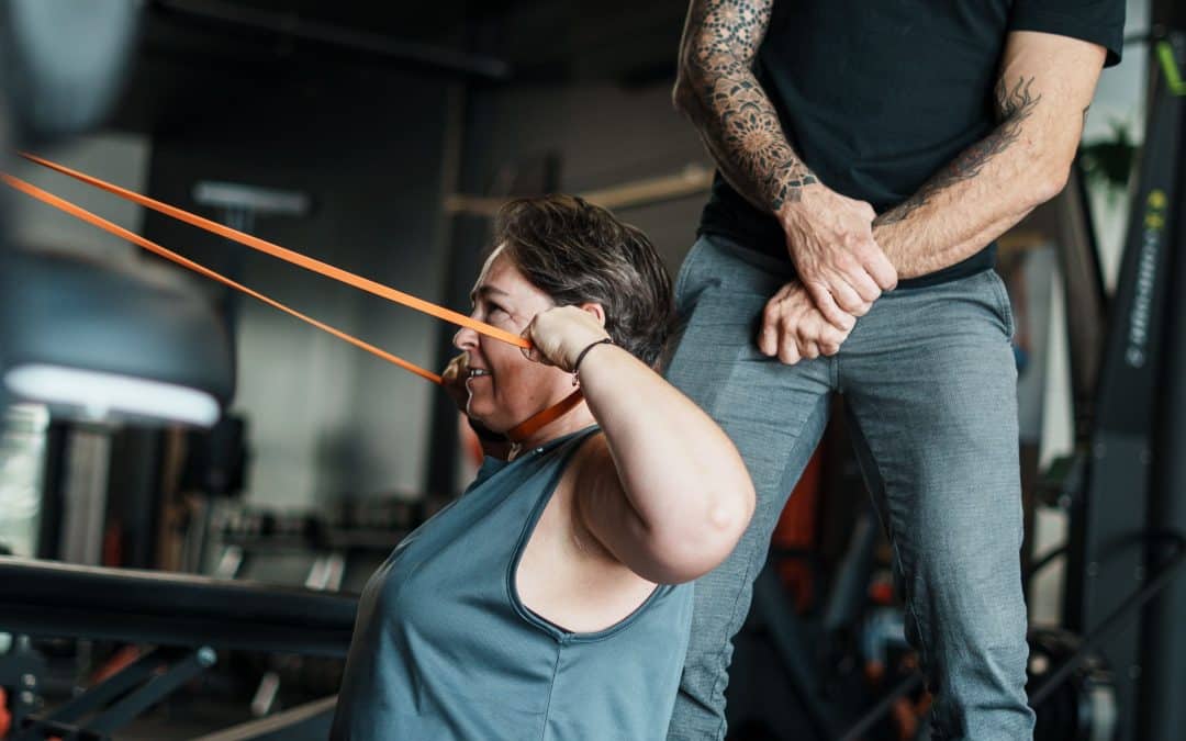 Cliente réalisant un tirage à l'élastique vers le visage avec l'aide d'un coach, un exercice qui renforce le haut du dos et soulage la nuque